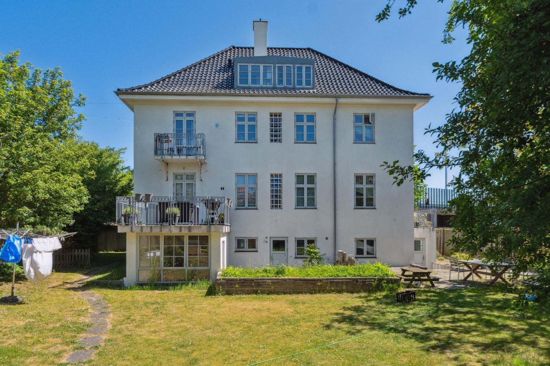 Hvidt kontorhuse med terrasse og have, lyse kontorlokaler, to solbrændestole og et bord på terrassen, trægrene og grønne