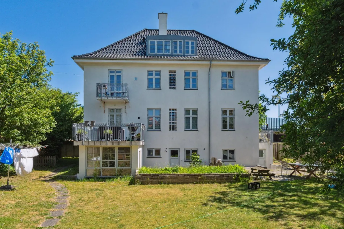 Hvidt kontorhuse med terrasse og have, lyse kontorlokaler, to solbrændestole og et bord på terrassen, trægrene og grønne