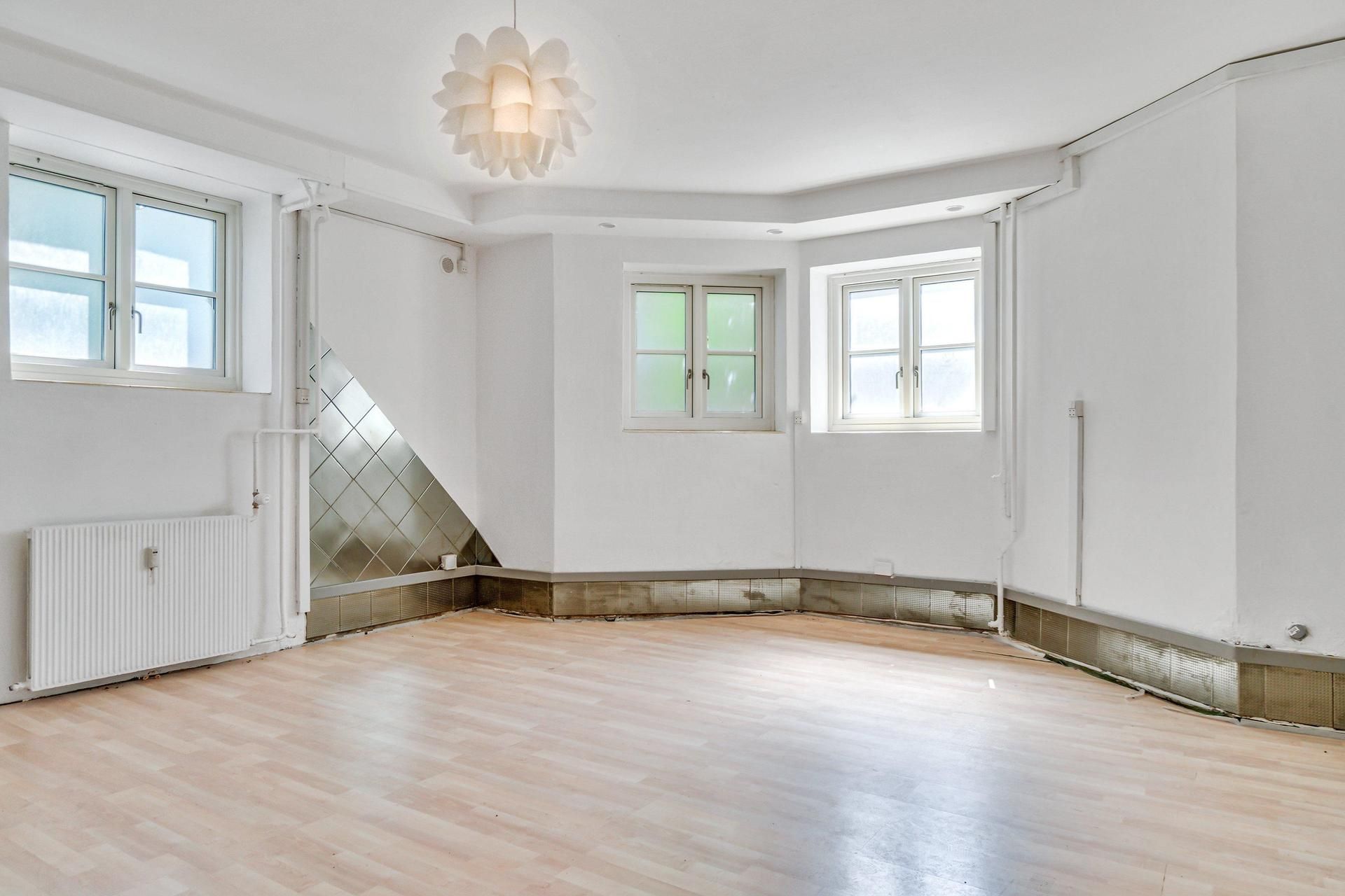 Et tomt kontor med hvide vægge og loft, trægulv, to vinduer og et lysekron.