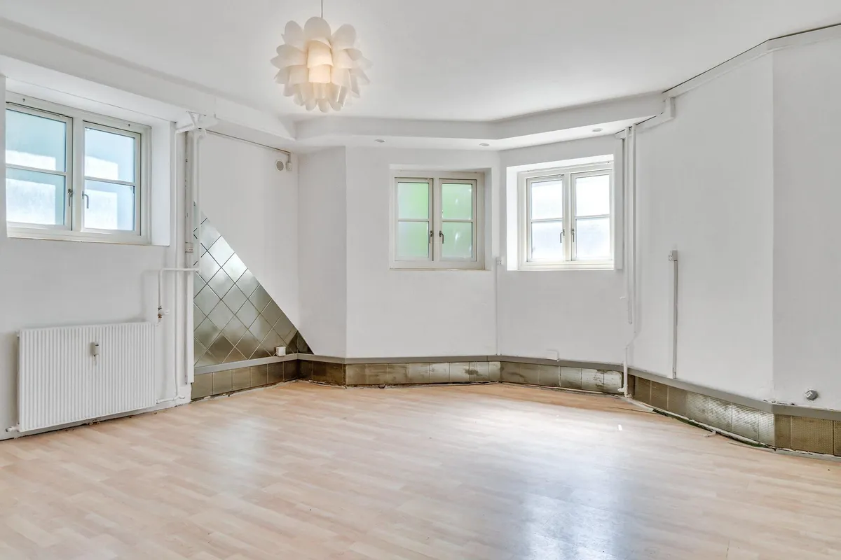 Et tomt kontor med hvide vægge og loft, trægulv, to vinduer og et lysekron.