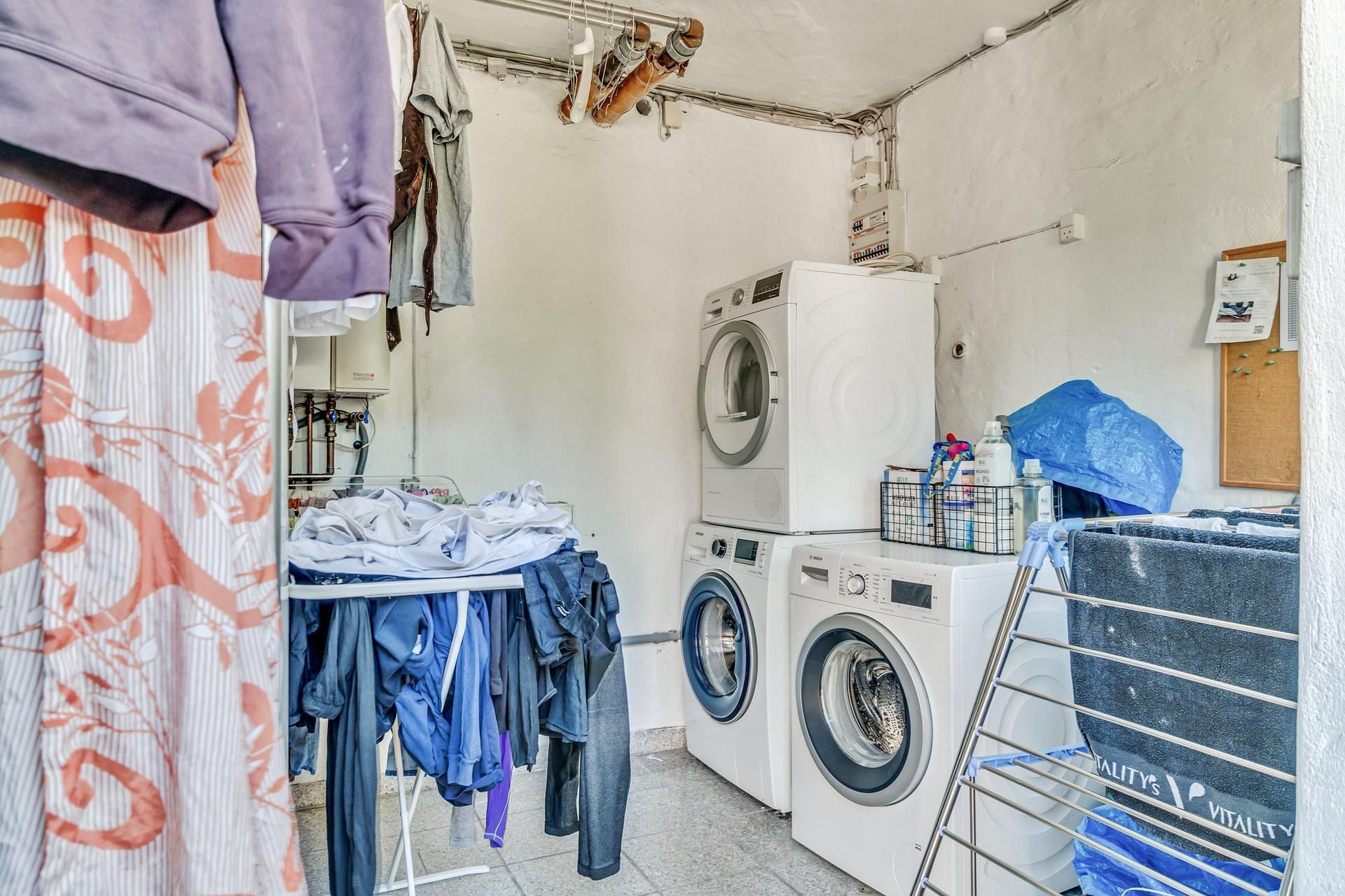 To hvide tøjvaskere står i et lille vaskeri med mange tøj på vasketøjshangere og en bøjlehylde med håndklæder