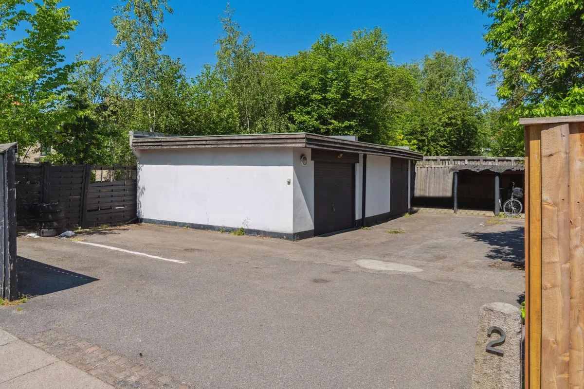 En stor parkeringsplads med to garager og en cykel parkeret udenfor. Garagedøre og lysarmatur er synlige. Bagved er der en