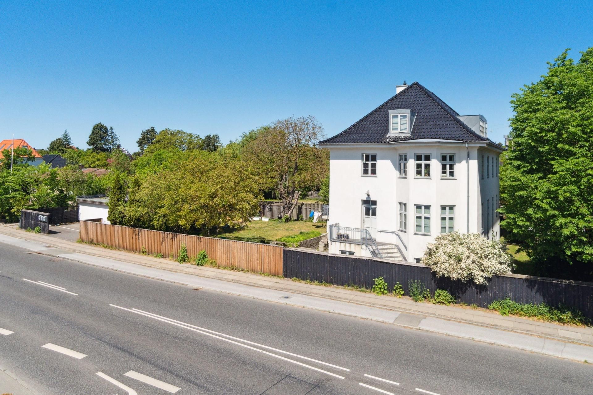 Lyst og stort lejlighedshus i Hellerup med have og garage. Høje huse i baggrunden og vej med biler. Træ- og metalhegn langs