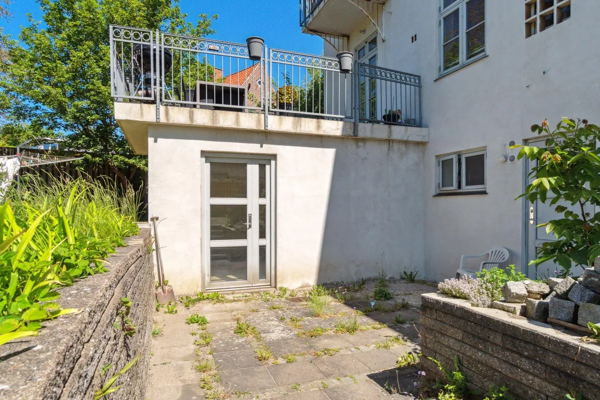 Kontor med terrasse og glasdør til gaden, skur med blomster og sten samt grønne planter langs mur