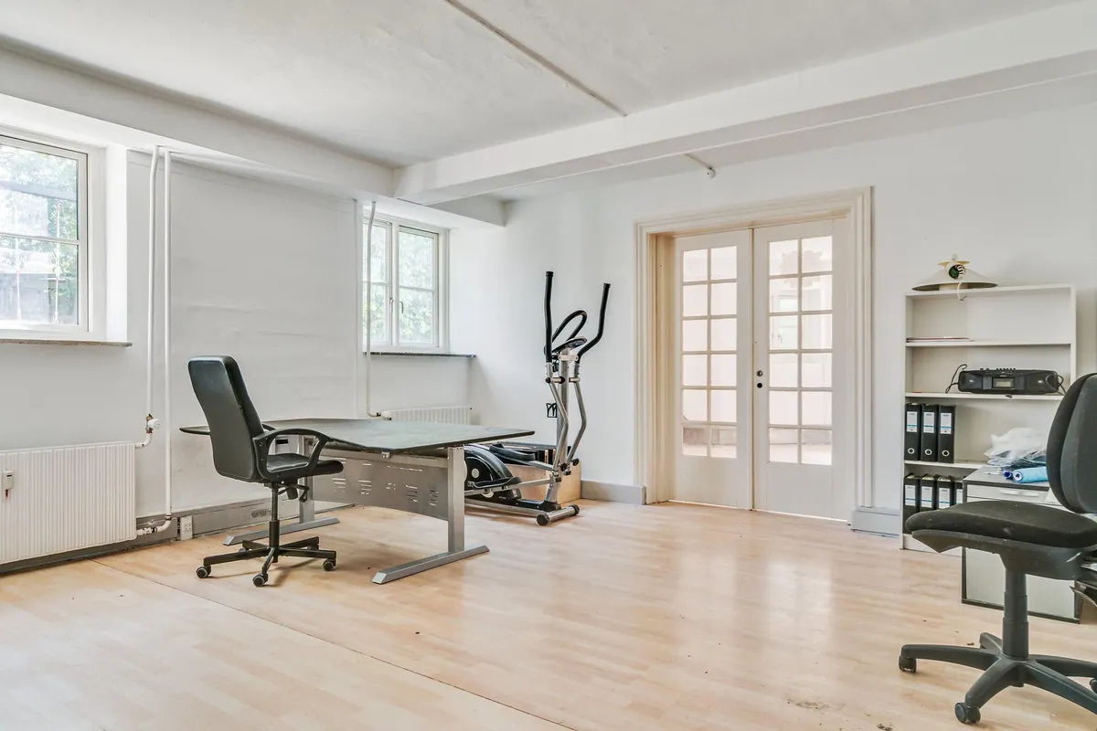 Kontor med skrivebord, trægulv, to lænestol, motionscykel, højre hjørne med hylde og glasdøre samt vinduer med udsigt til