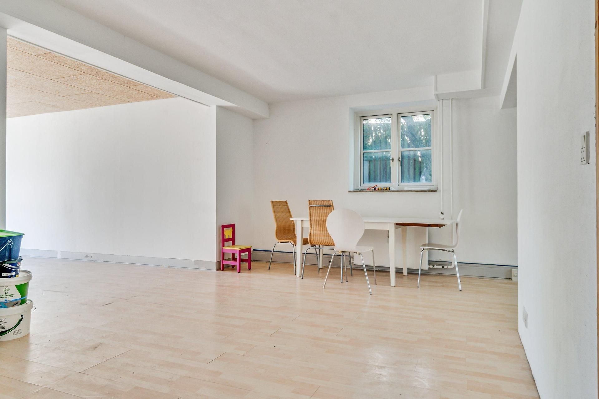 Hvidt lokale med trægulv og tre hvide stole samt en lille pink og hvid stol med skriftstykke på bordet