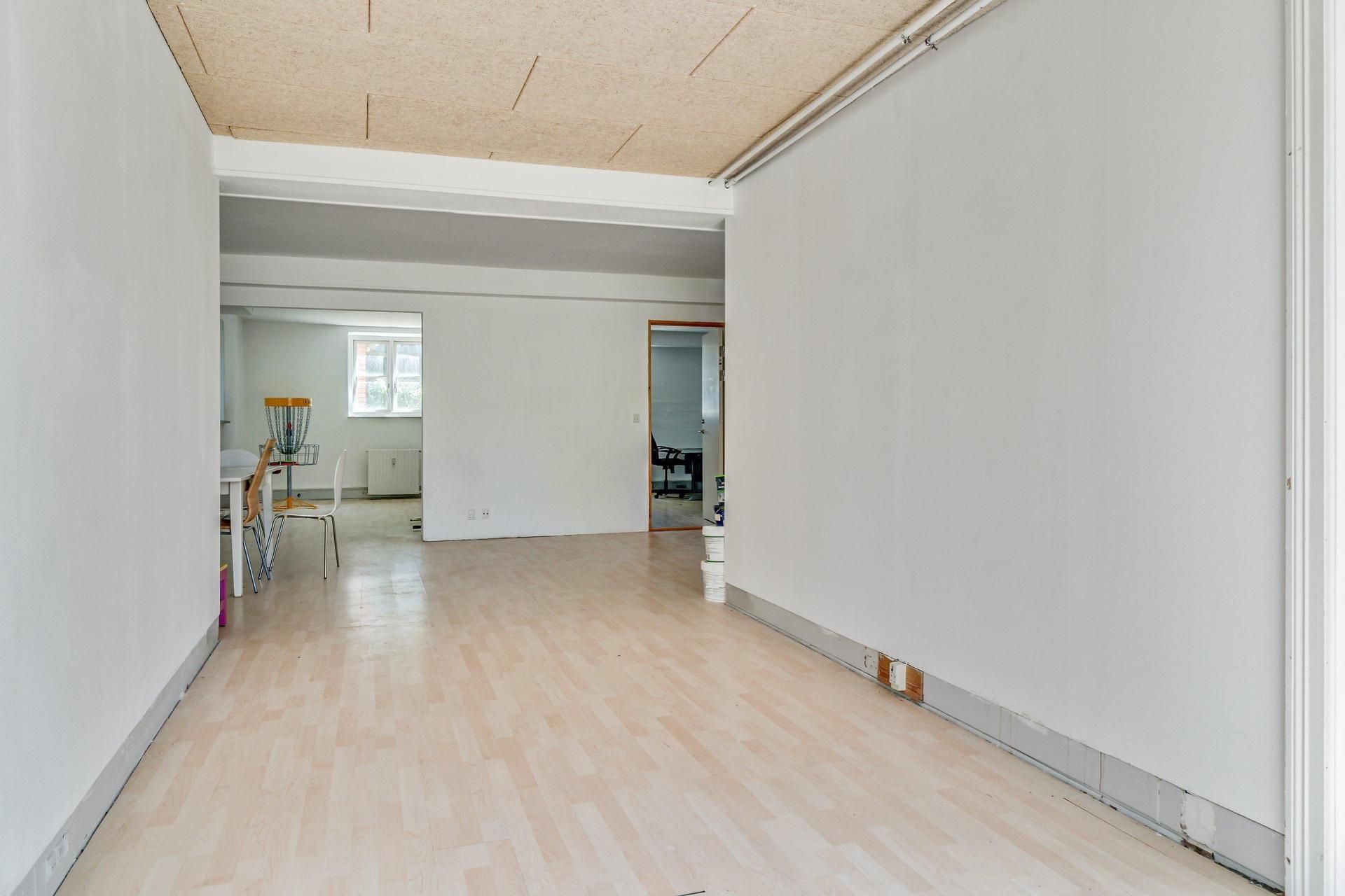 Kontorlokaler med lyse vægge og trægulv samt møbler som bord og stole i enden af rummet og et vindue med udsigt til en anden