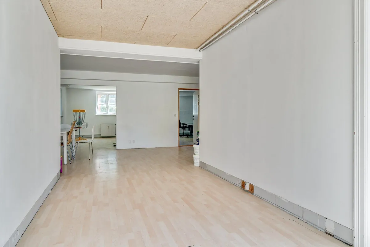 Kontorlokaler med lyse vægge og trægulv samt møbler som bord og stole i enden af rummet og et vindue med udsigt til en anden