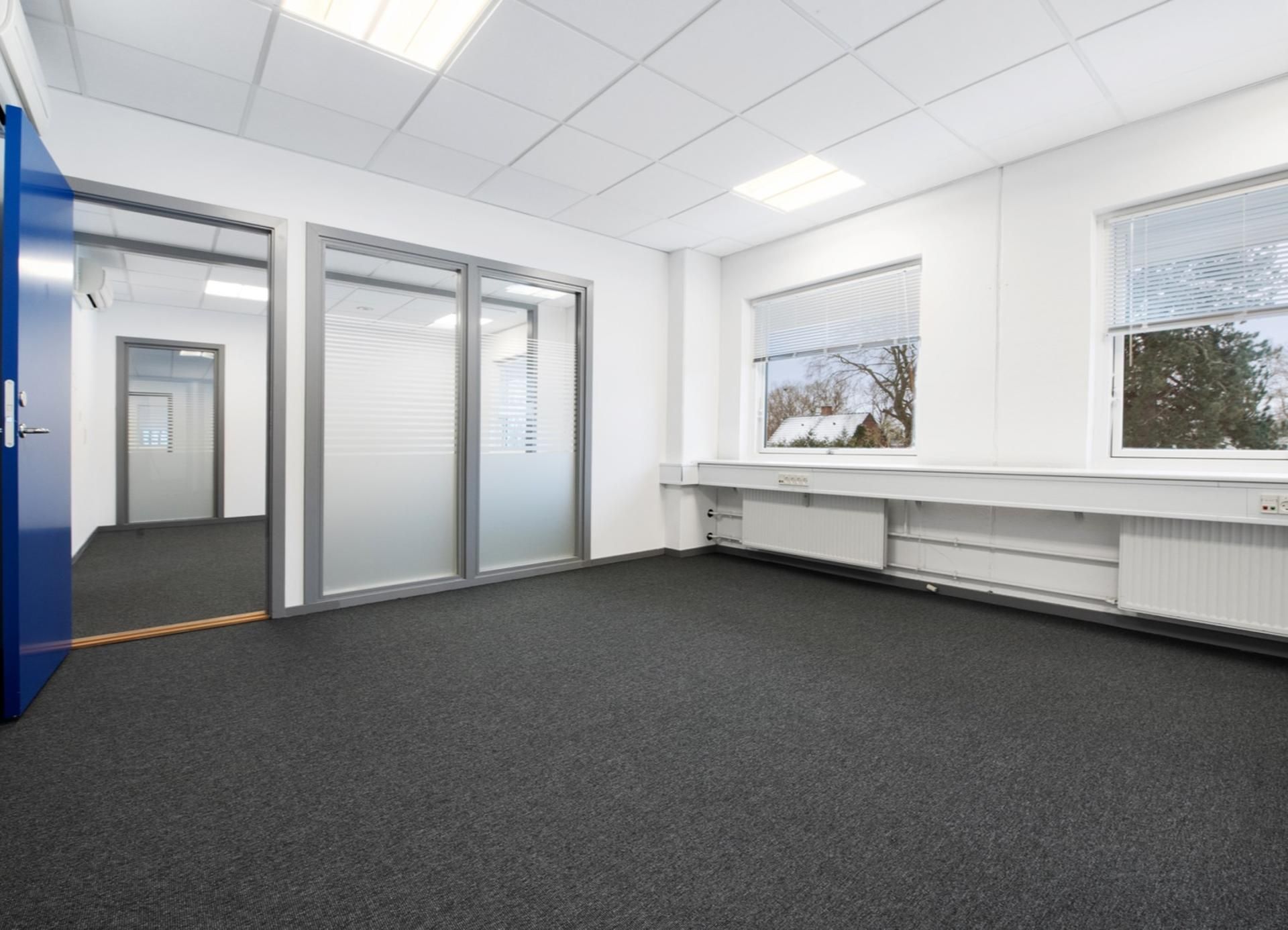 Store, tomme kontorlokaler med lyse vægge, hvide lofter, tætte ruder og blå døre samt lysegule gulvbelægning og møbler, der