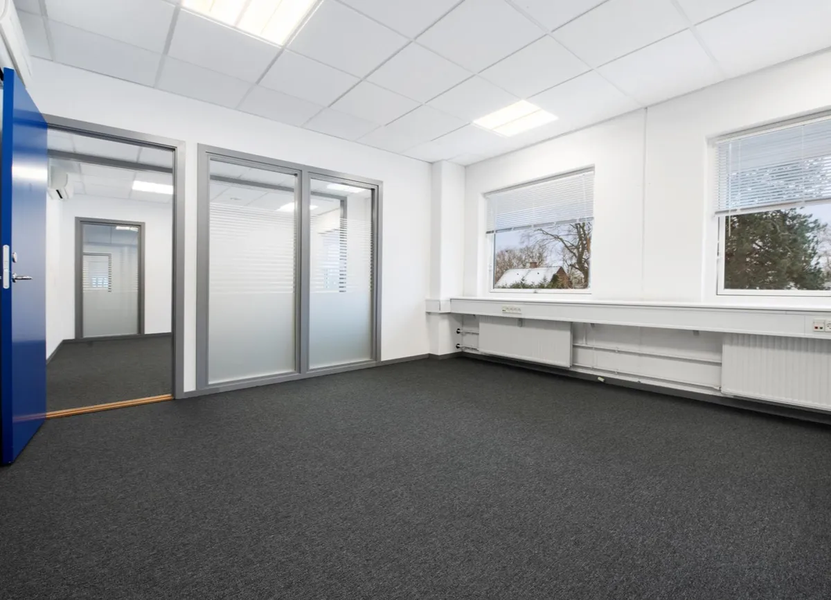 Store, tomme kontorlokaler med lyse vægge, hvide lofter, tætte ruder og blå døre samt lysegule gulvbelægning og møbler, der