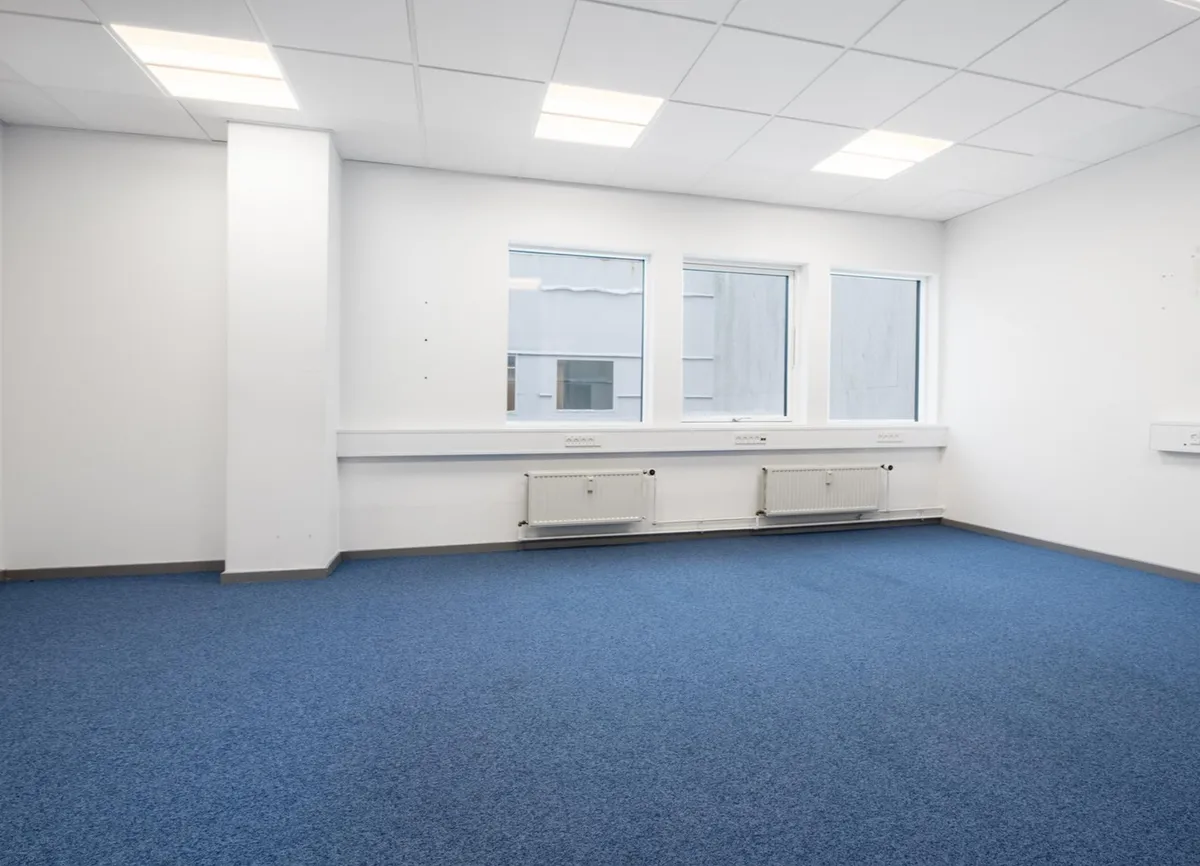 Kontorlokaler med hvidt vægmaleri og blå gulvbelægning samt tre store ruder og hvide radiatorer langs væggen