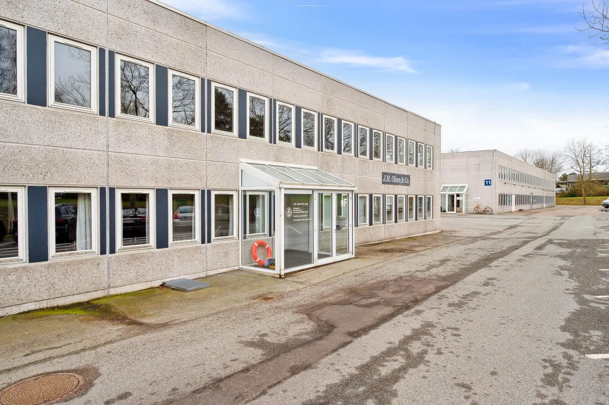 Kontorbyggeri med store ruder og glasdør til indgang. Parkeringsplads med skrald og en cykel parkeret. Bygning med skilt,