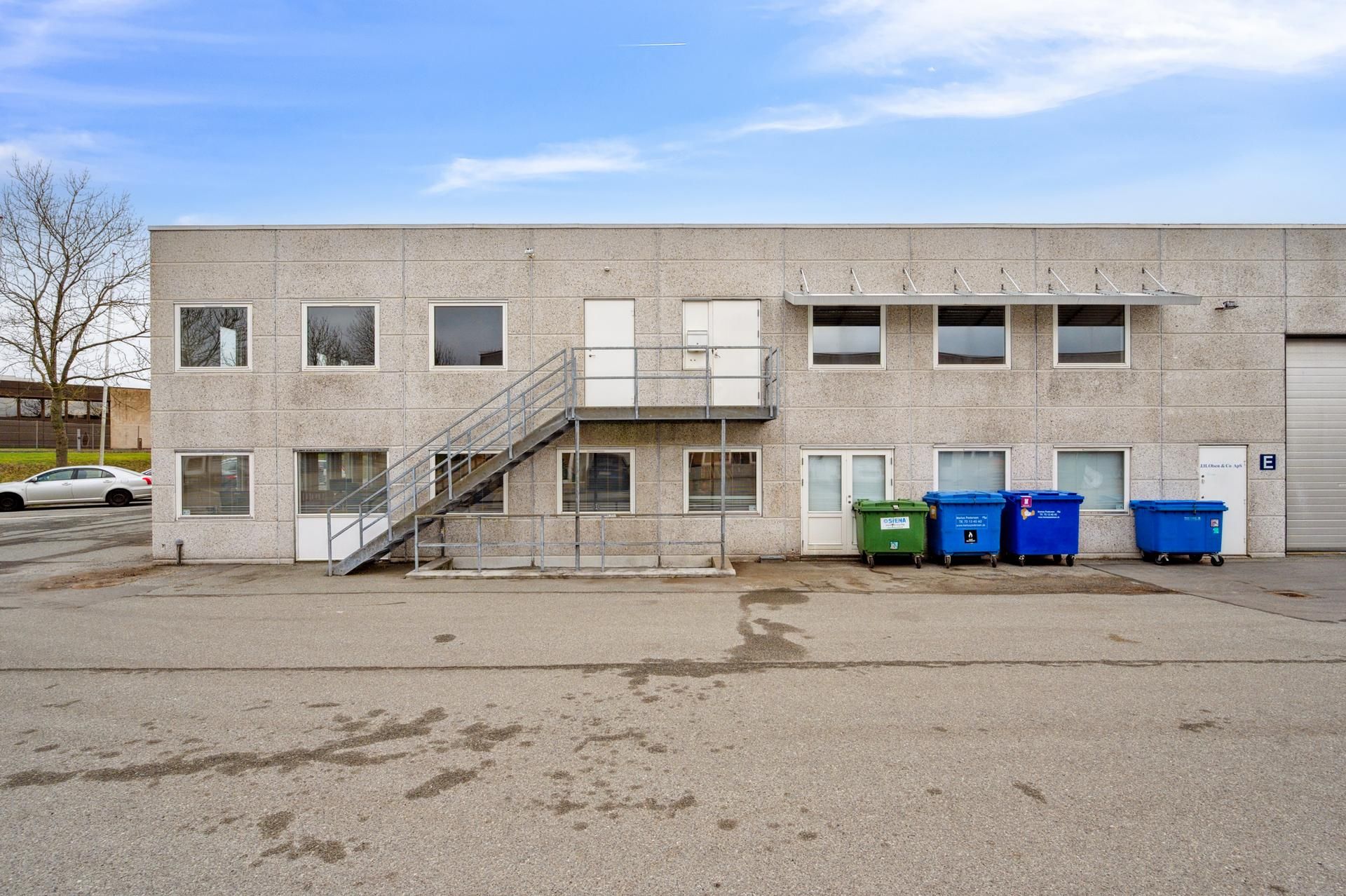 Foran et moderne kontorbyggeri er der parkeringsplads, tre stålkasser og et trappeforløb med metalgelænder