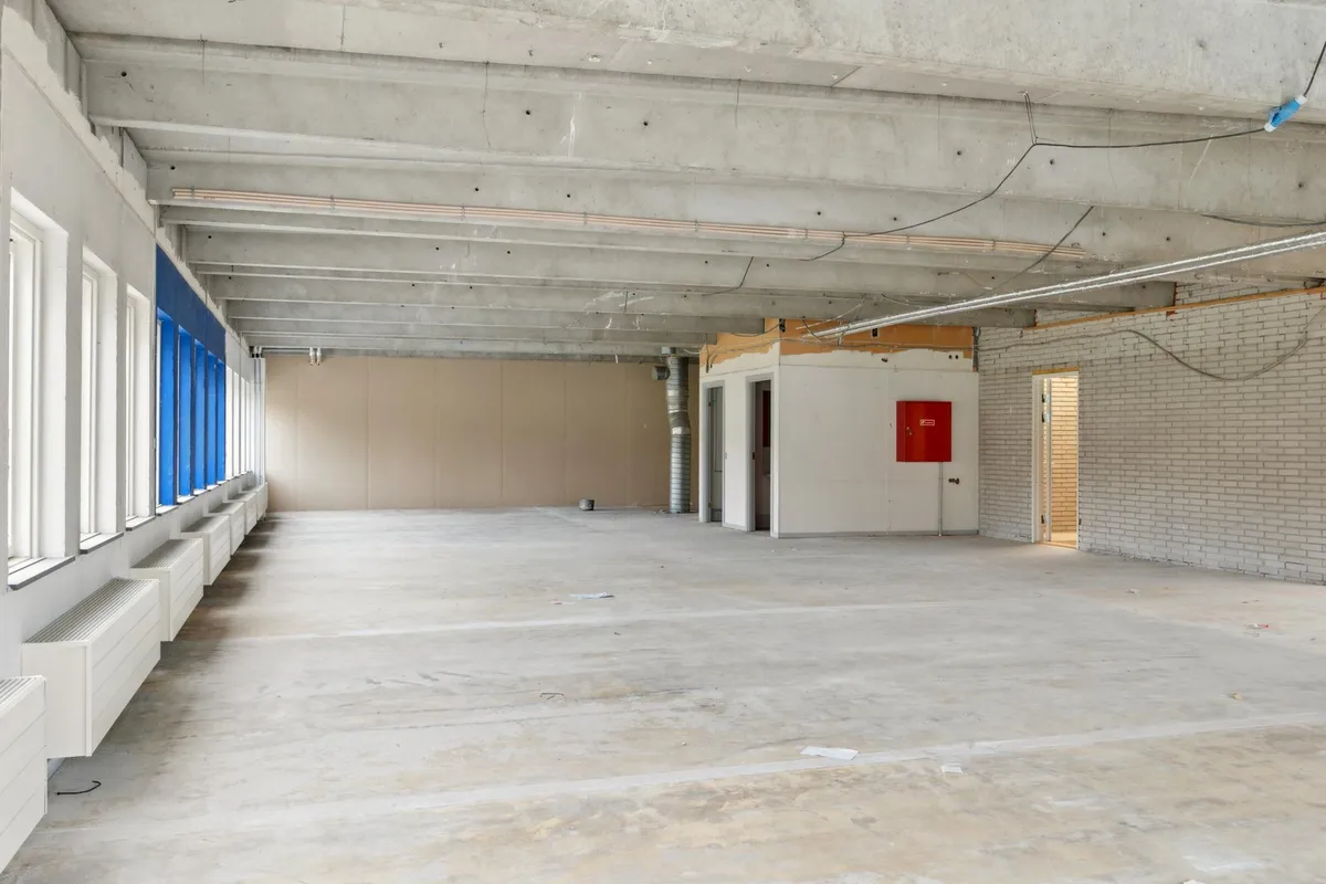 Indenfor en stor, tom bygning med høje lofter, hvide vægge og en gulvbelægning af beton. Der er flere hvide radiatorer langs