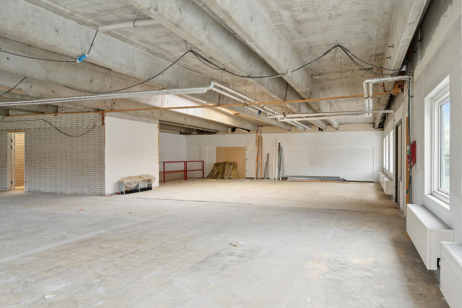 Lokal i Skovlunde med ubenyttet rum, skraldecontainer, røde metalbjælker, vinduer og radiatorer samt hængende rørværk og