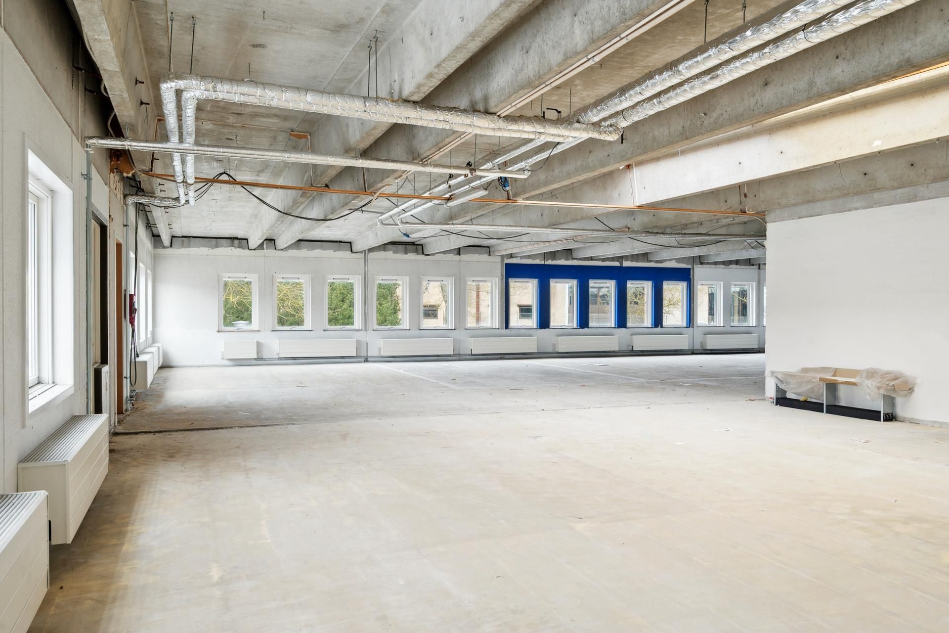 Et tomt lokale med hvide vægge og cementgulve, lysevinduer og luftkanaler og piper i loftet, samt en træbord med