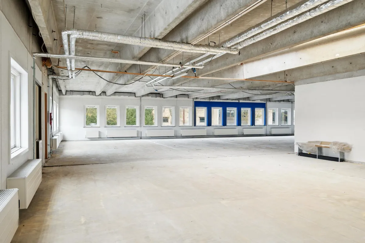 Et tomt lokale med hvide vægge og cementgulve, lysevinduer og luftkanaler og piper i loftet, samt en træbord med