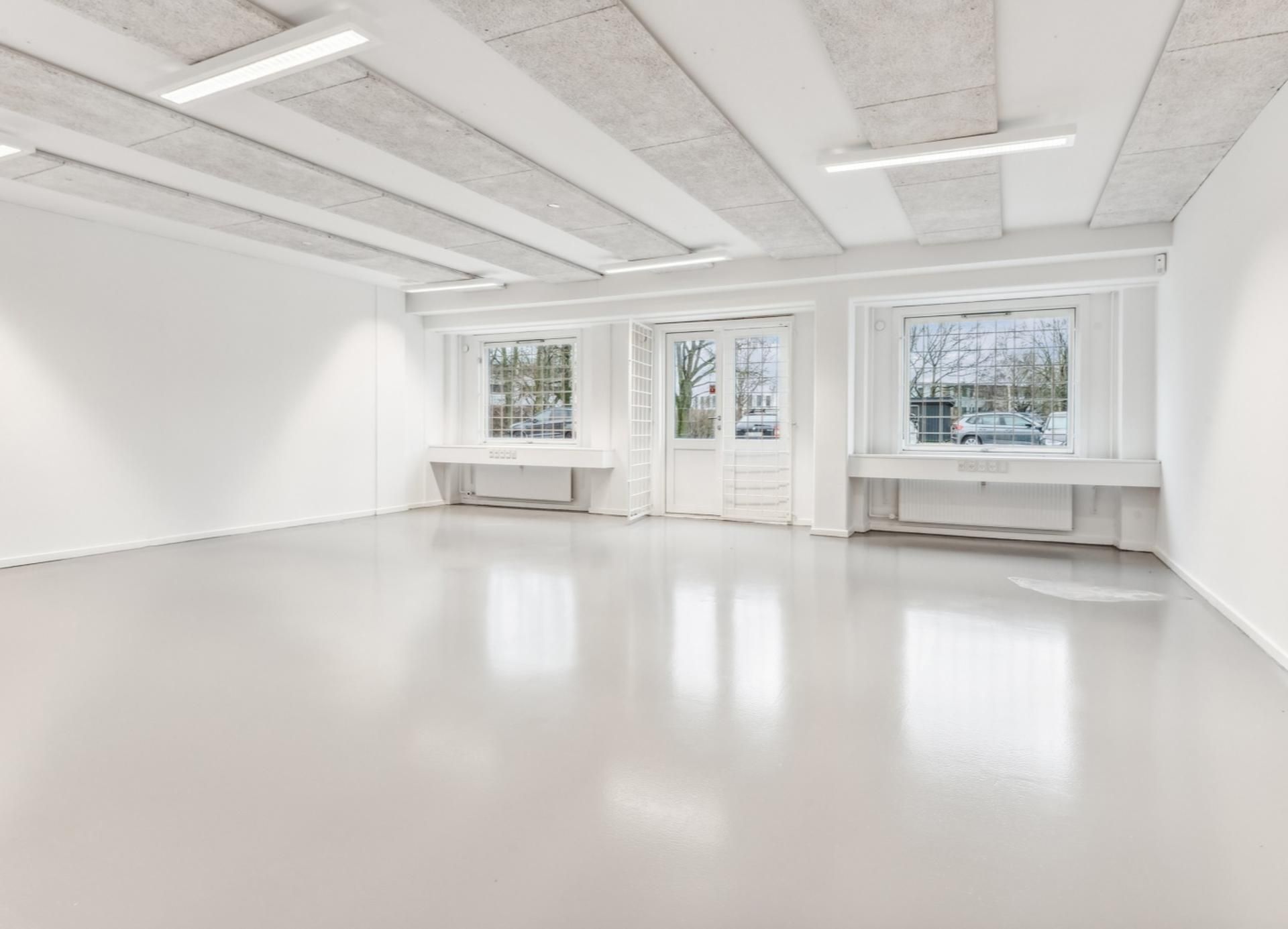 Ledig erhvervslejemål med lyse lofter og store vinduer med udsigt til parkeringsplads og biler i baggrunden