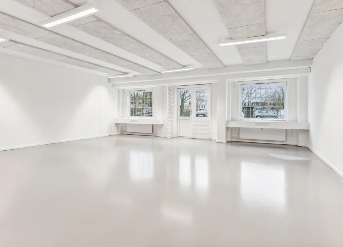 Ledig erhvervslejemål med lyse lofter og store vinduer med udsigt til parkeringsplads og biler i baggrunden