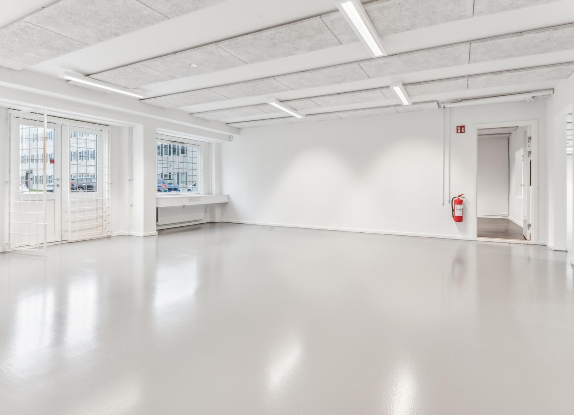 Et værelse med hvide vægge, højt loft, lyskilder, en dør med rødt brandslukker og vinduer med udsigt til parkeringsplads