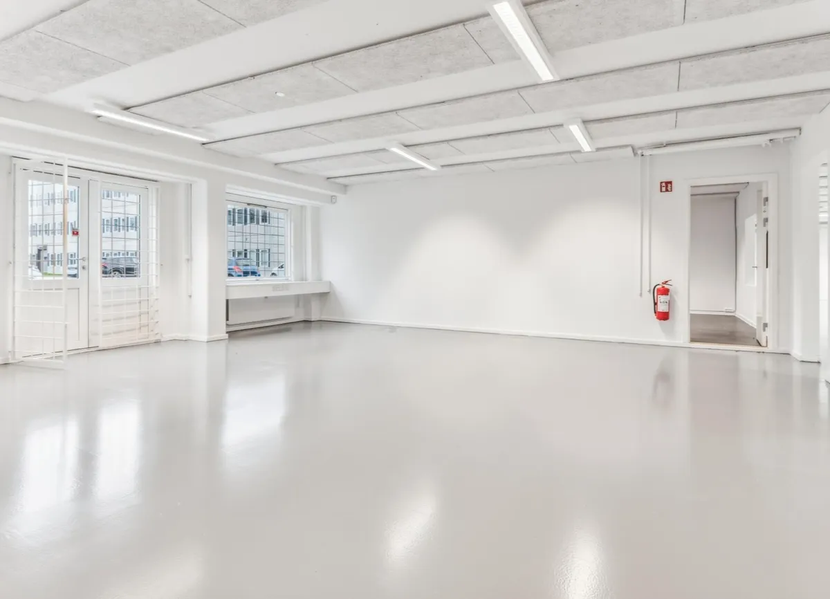 Et værelse med hvide vægge, højt loft, lyskilder, en dør med rødt brandslukker og vinduer med udsigt til parkeringsplads