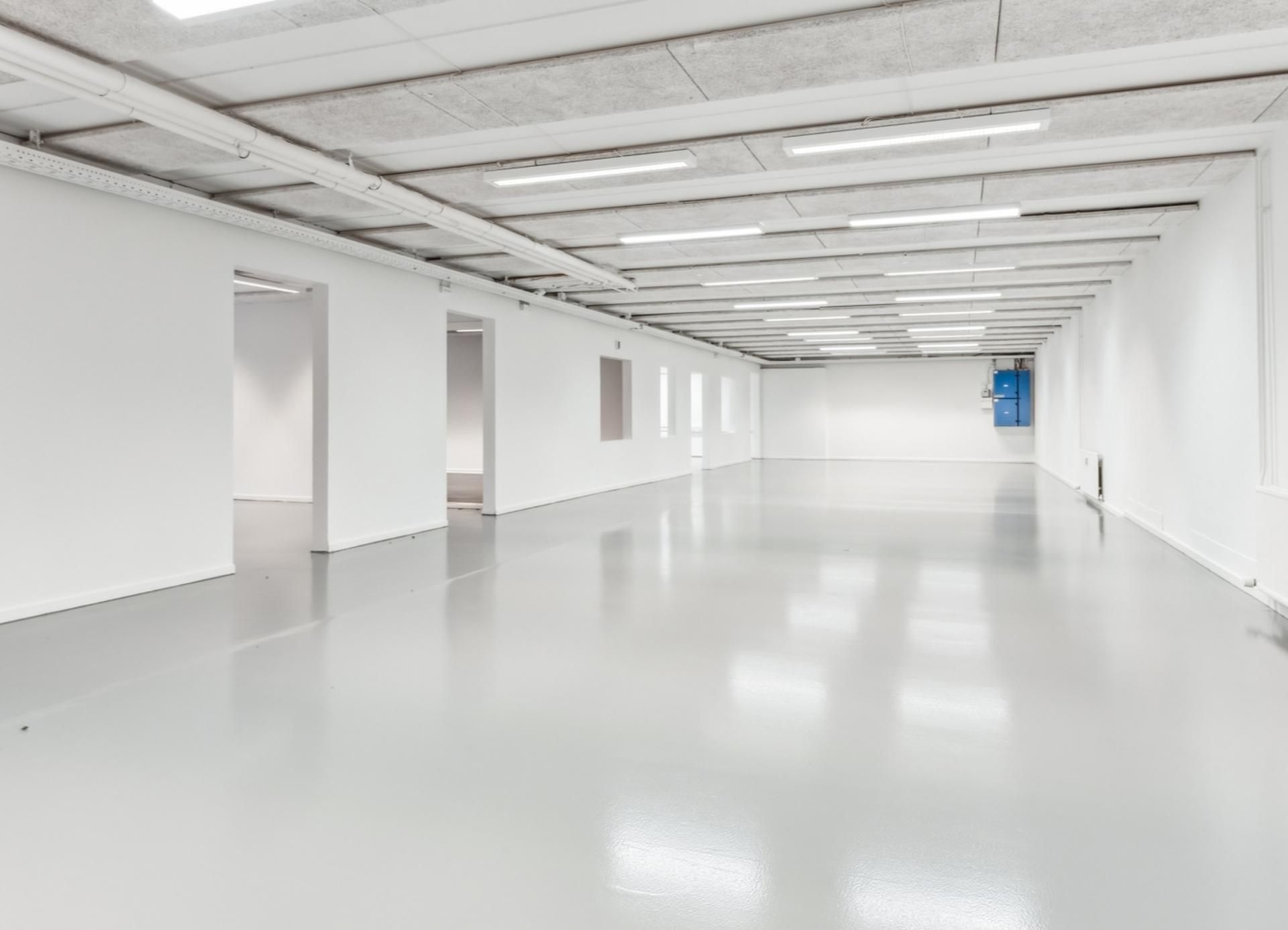 En lang, tom gang med hvide vægge, gulve og loft, højt, funktionelt lys og åbninger mod siderne