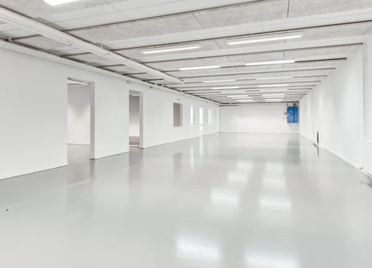 En lang, tom gang med hvide vægge, gulve og loft, højt, funktionelt lys og åbninger mod siderne