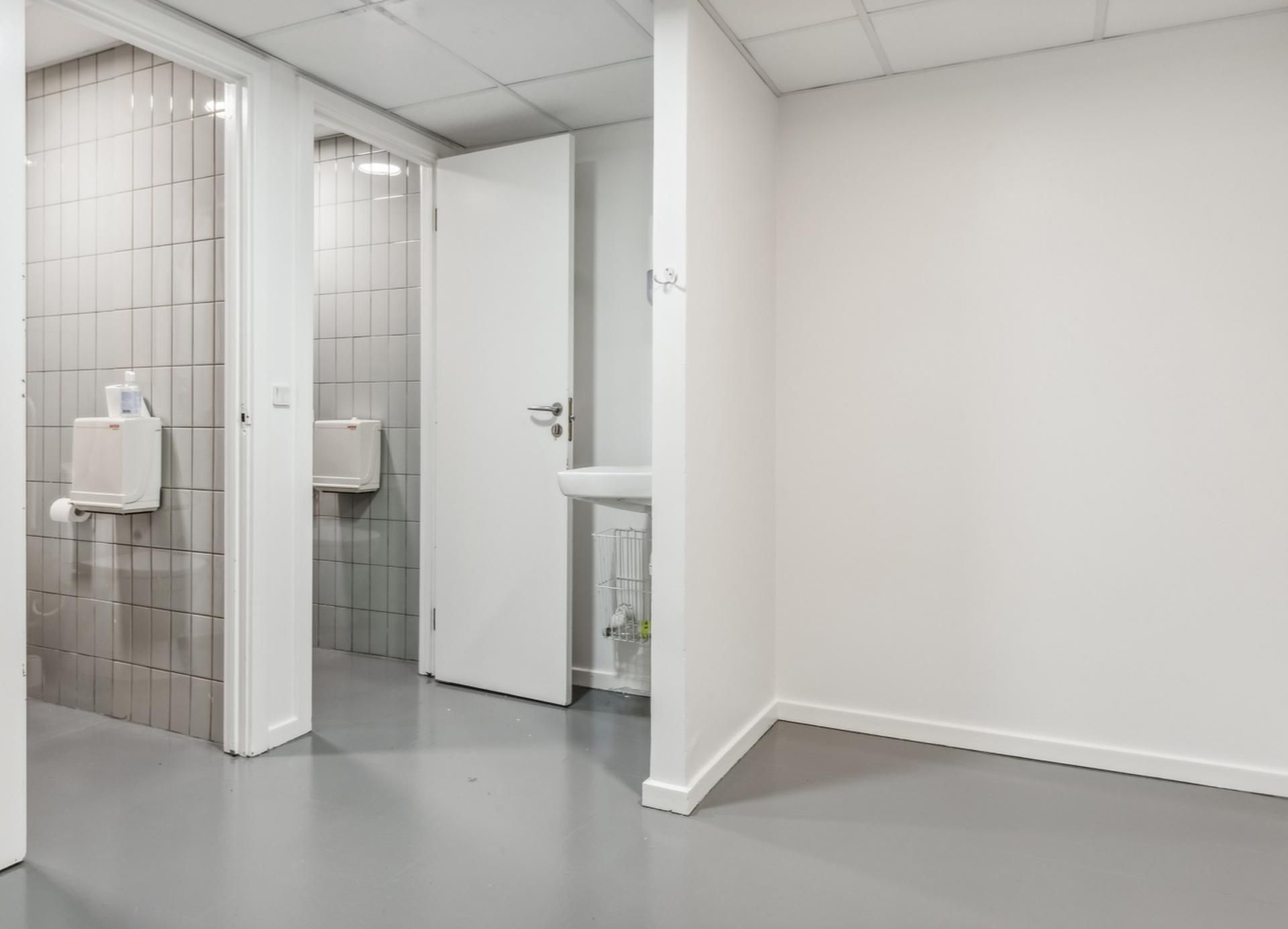 Toilette med hvidt loft og gulv, lukket dør og hvidt vægforløb, to toiletter og et hvidt skarpt vask.