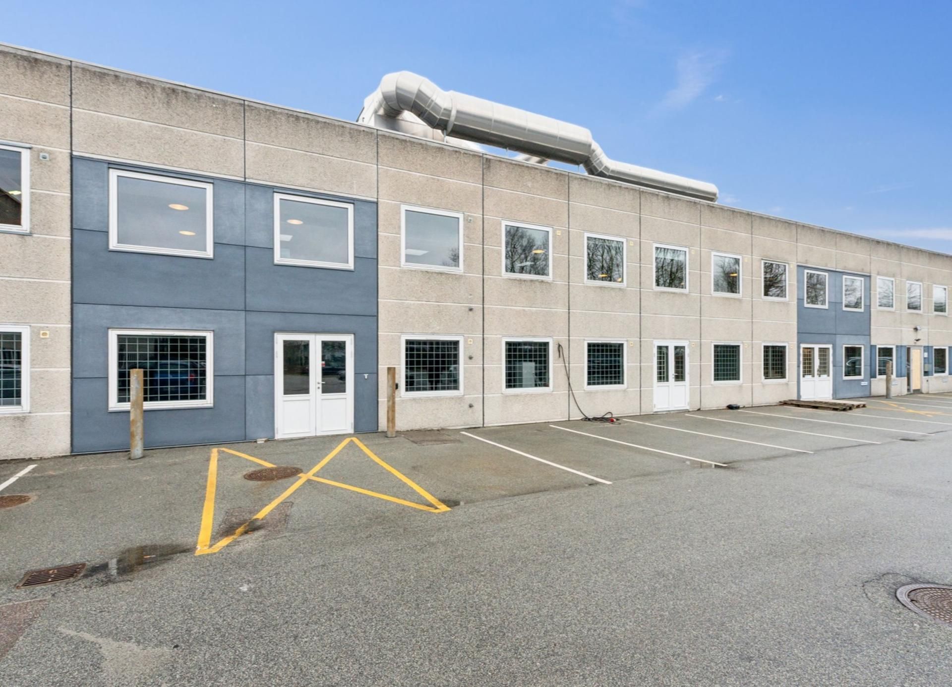 Store bygning med glasruder og døre, hvorfra lys skinner ind, og der er parkeringsplads med gule linjer og en stor skorsten