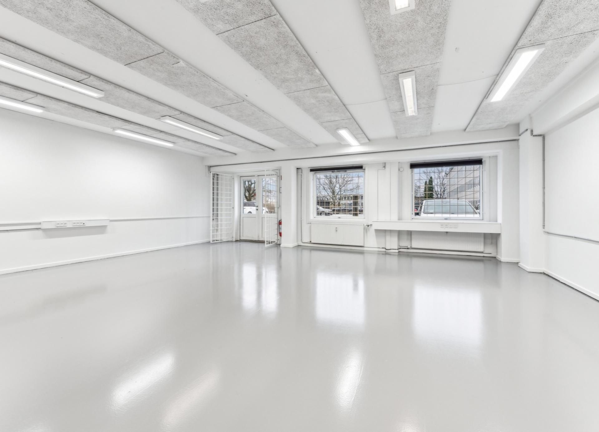 En tom industrilokal med hvide vægge og gulve samt højt, lysekældret loft med brændenælde mønstre og fire store vinduer