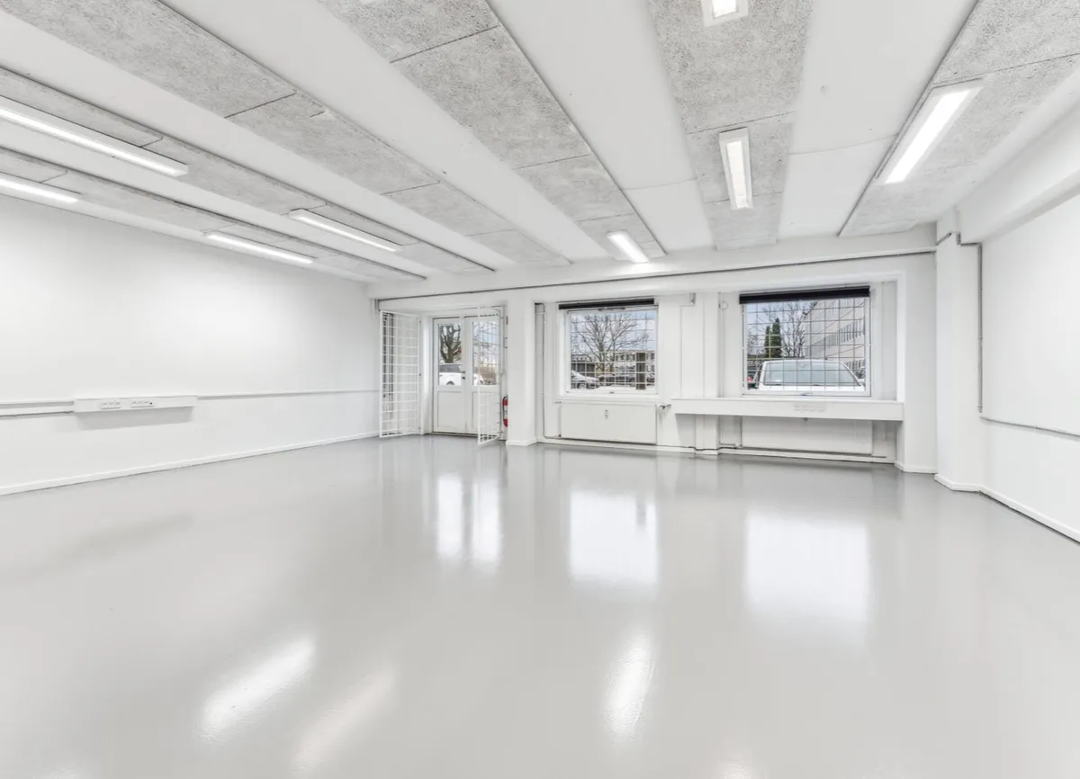 En tom industrilokal med hvide vægge og gulve samt højt, lysekældret loft med brændenælde mønstre og fire store vinduer