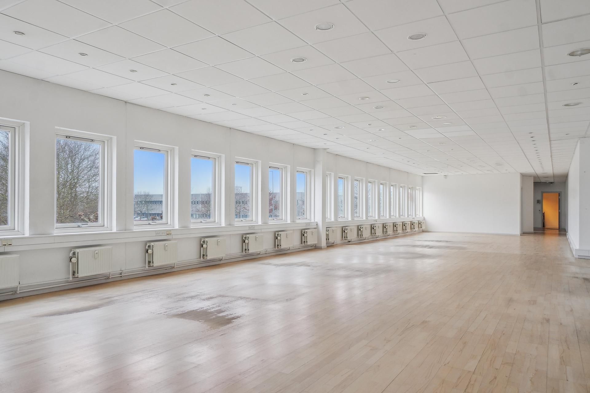 Lyst og velholdt kontor med trægulv og hvide vægge, 12 store vinduer og 10 radiatorer langs væggen, dør til siderum