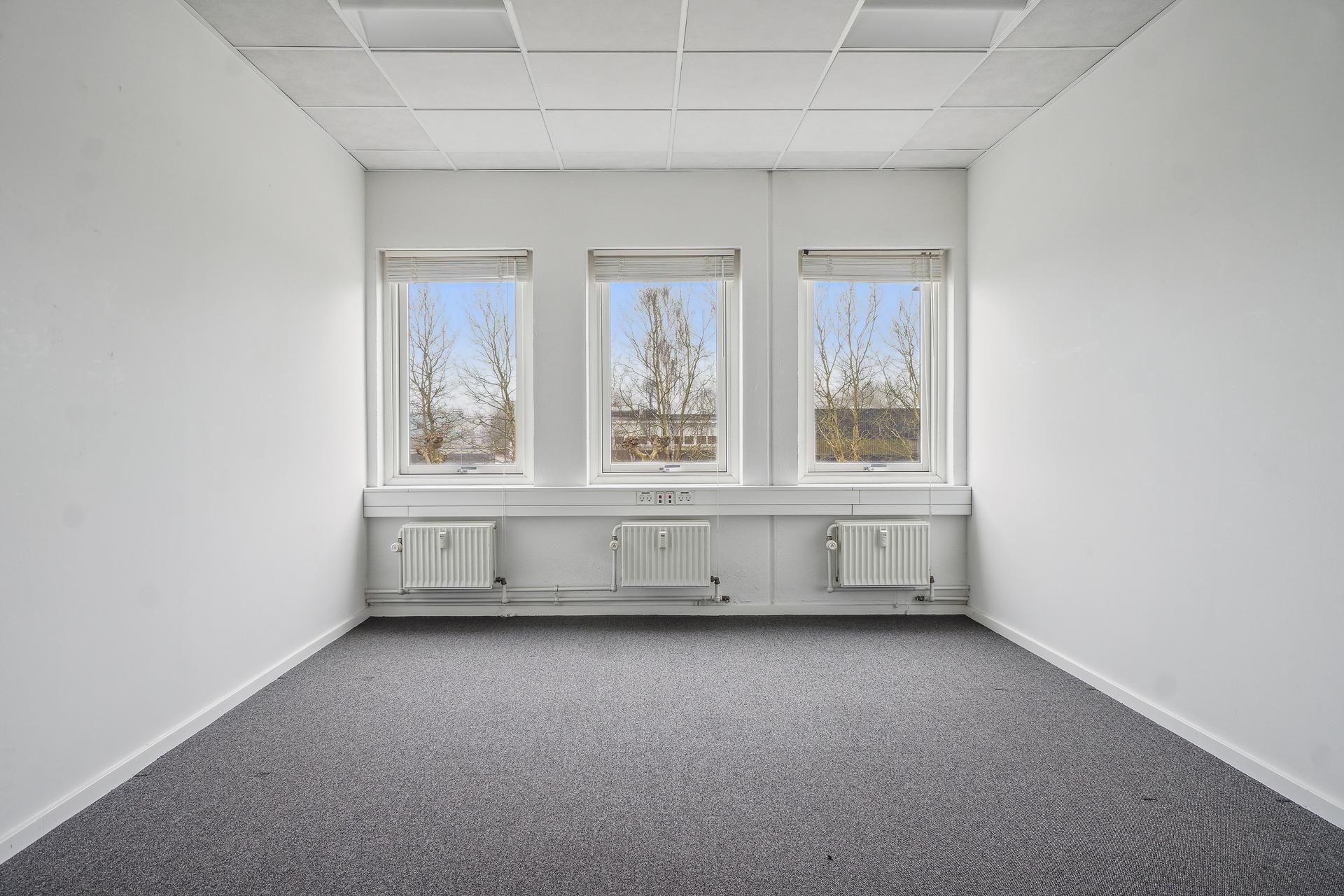 Et tomt kontorlokal med tre vinduer, hvide vægge og loft, gulvbelægning og tre radiatorer i bunden af væggen og to