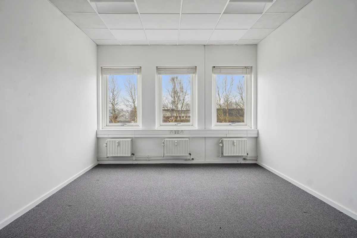 Et tomt kontorlokal med tre vinduer, hvide vægge og loft, gulvbelægning og tre radiatorer i bunden af væggen og to