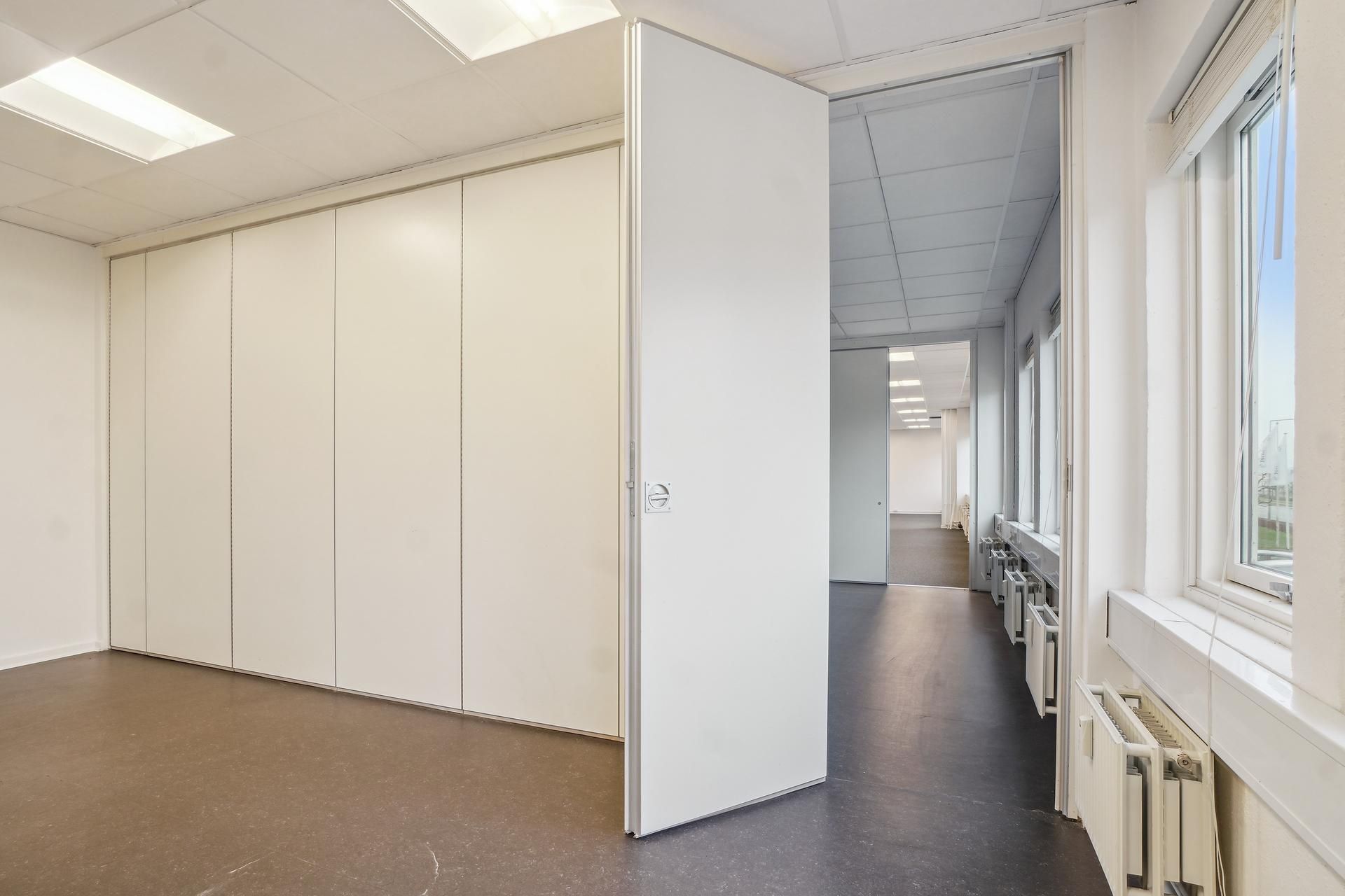 Et hvidt kontorlokal med åben højre sidedør og flere vægge med hvide skurpaneler. Væggen mod højre har et vinduesparti med
