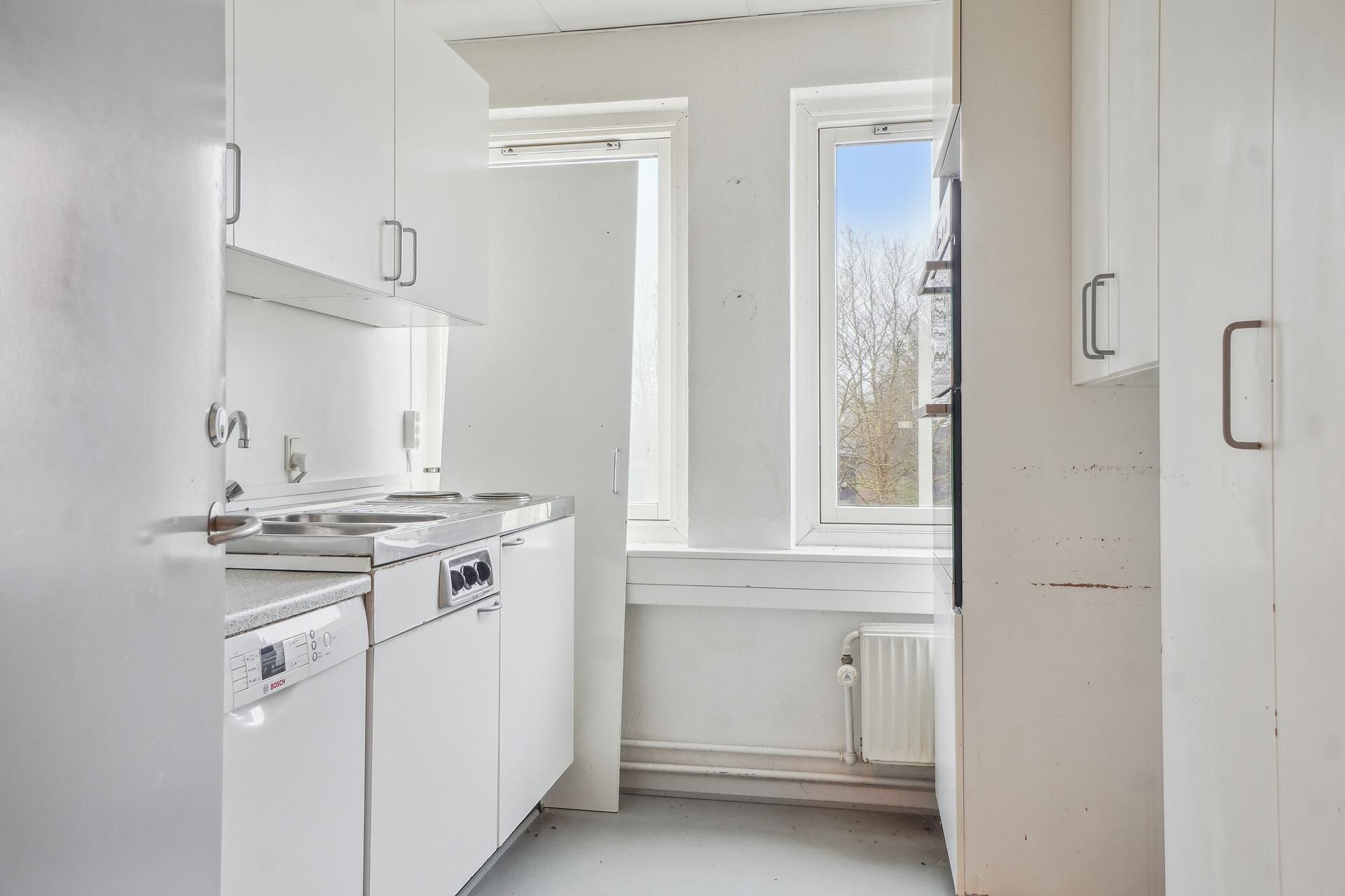 Et moderne og funktionelt køkken med hvide vægge og skabe, vinduer med gardiner, og en ren og klar udsigt udenfor