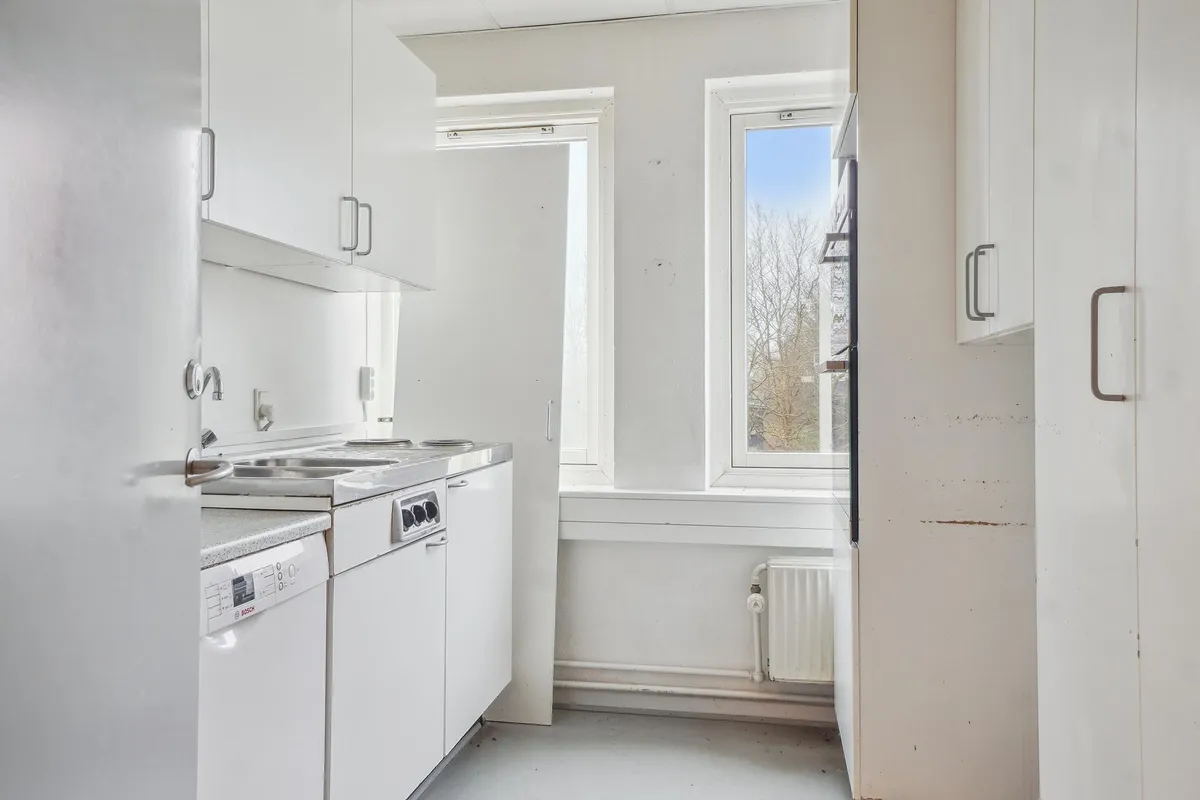 Et moderne og funktionelt køkken med hvide vægge og skabe, vinduer med gardiner, og en ren og klar udsigt udenfor