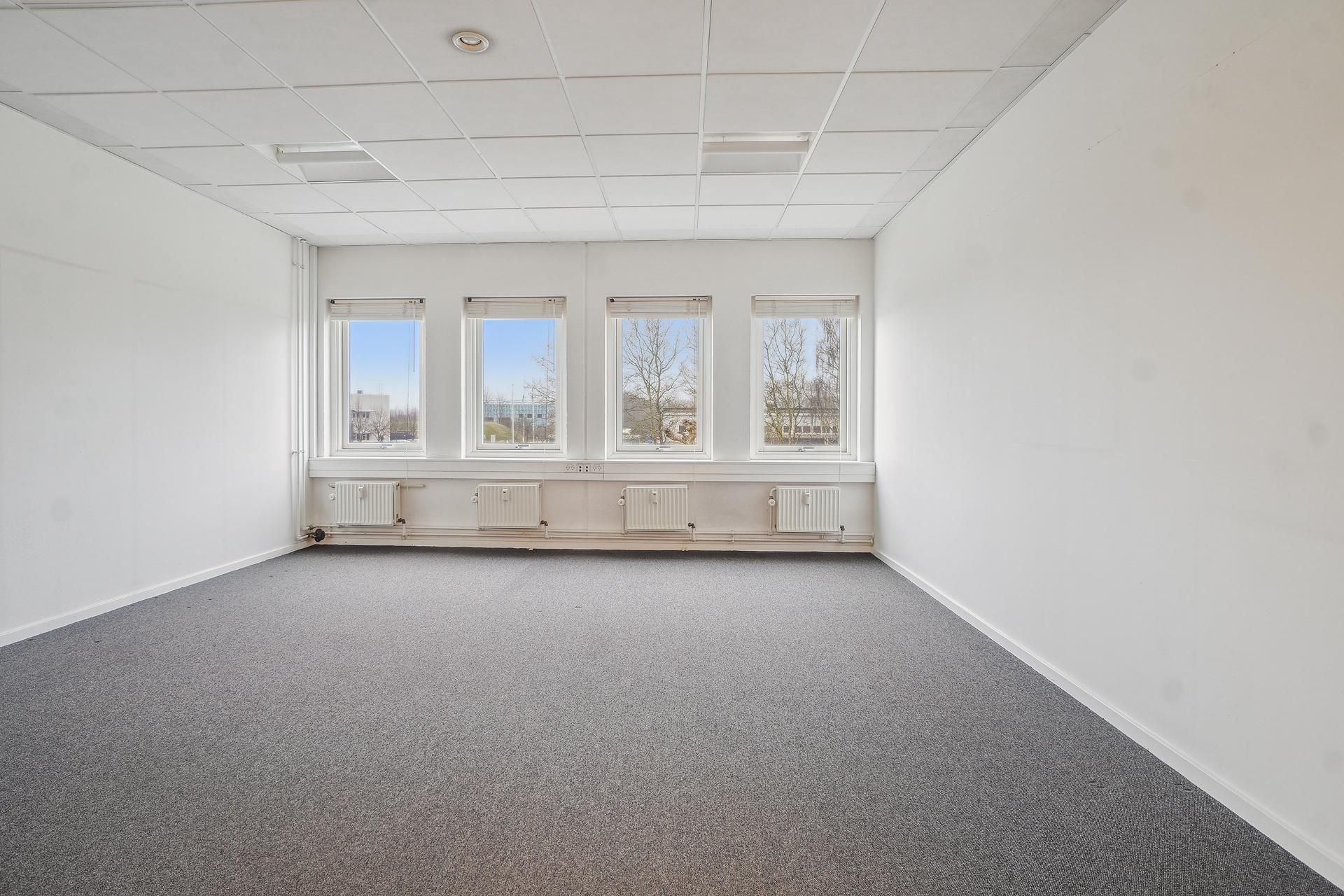 En tom kontor med tre vinduer og hvide vægge, gulv og loft. Værelset har en grå gulvbelægning og tre hvide radiatorer. Et