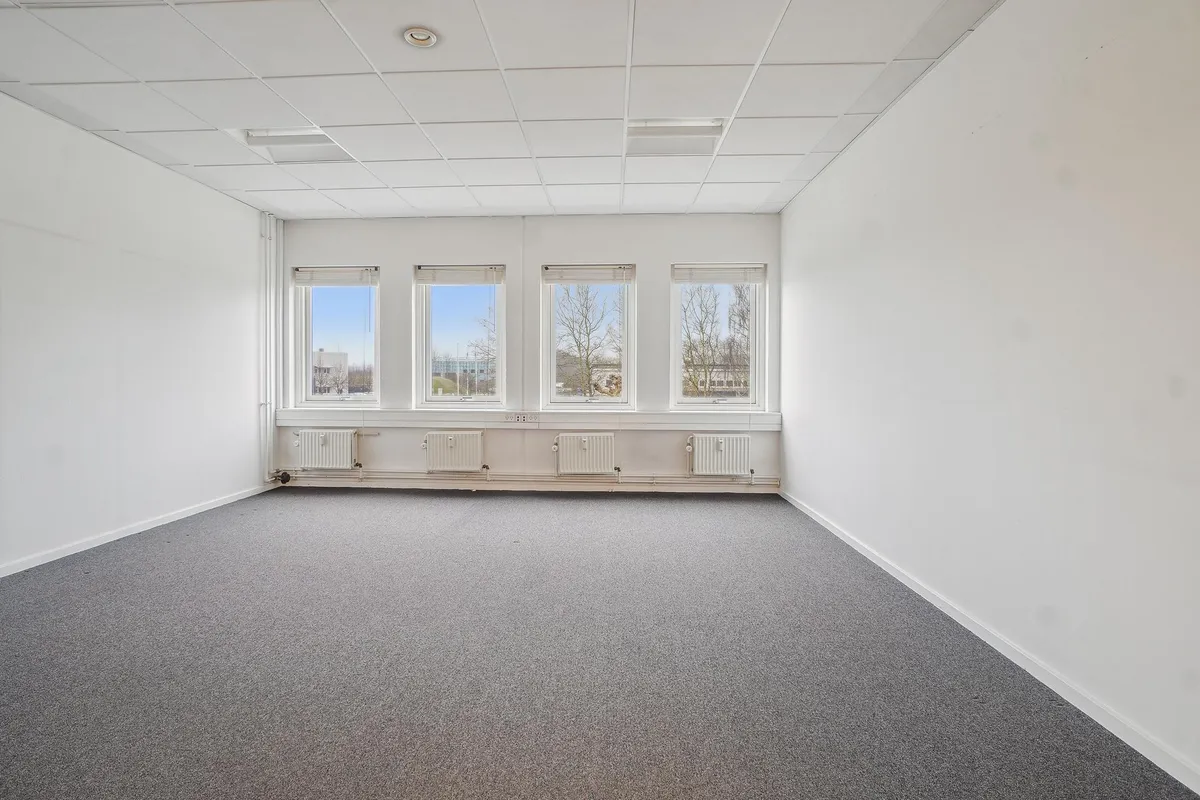 En tom kontor med tre vinduer og hvide vægge, gulv og loft. Værelset har en grå gulvbelægning og tre hvide radiatorer. Et