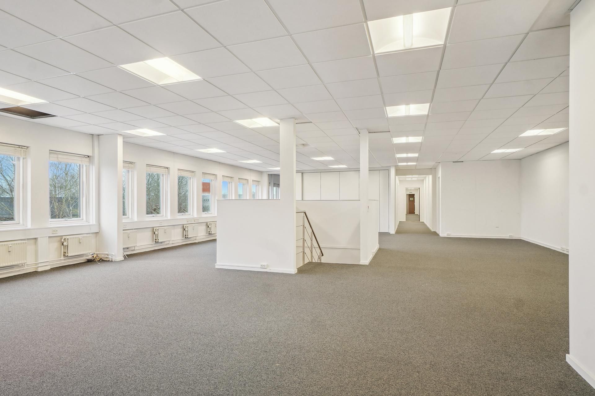 Et tomt og lyst kontorlokal med hvide vægge, gulvtæppe og vinduer med udsigter til træer udenfor.