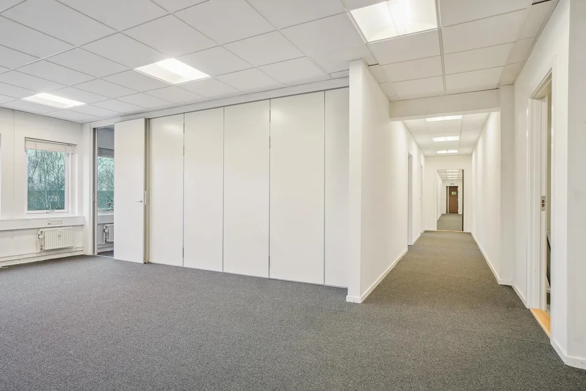 En lys, rummelig og velholdt kontorbygning med hvide vægge og gulv, hvor der er en dør med glas og et vindue med en jalusi.