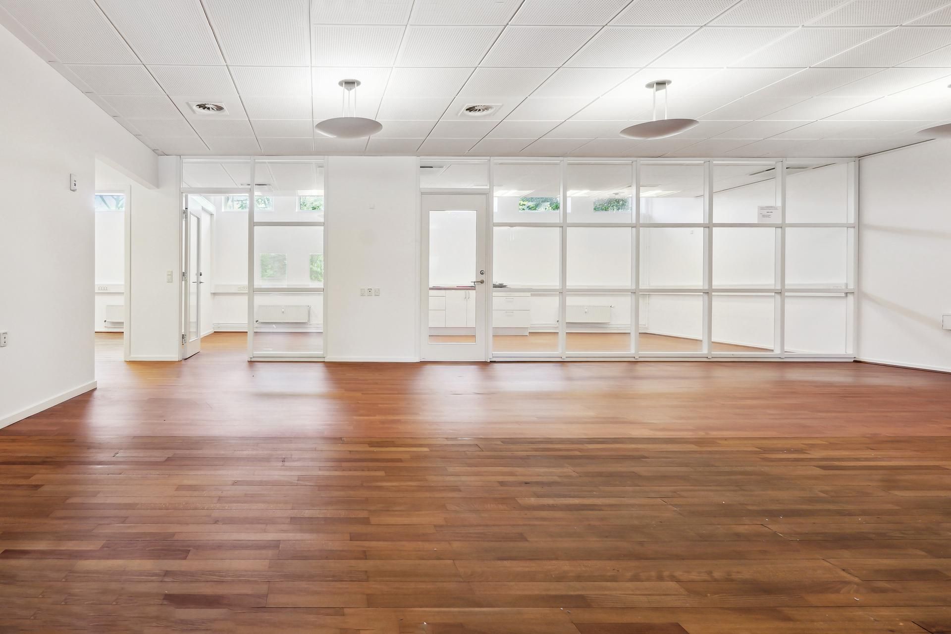 Kontorlokation med hvide vægge og lyse loftspaneler samt glasvinduer og trægulv
