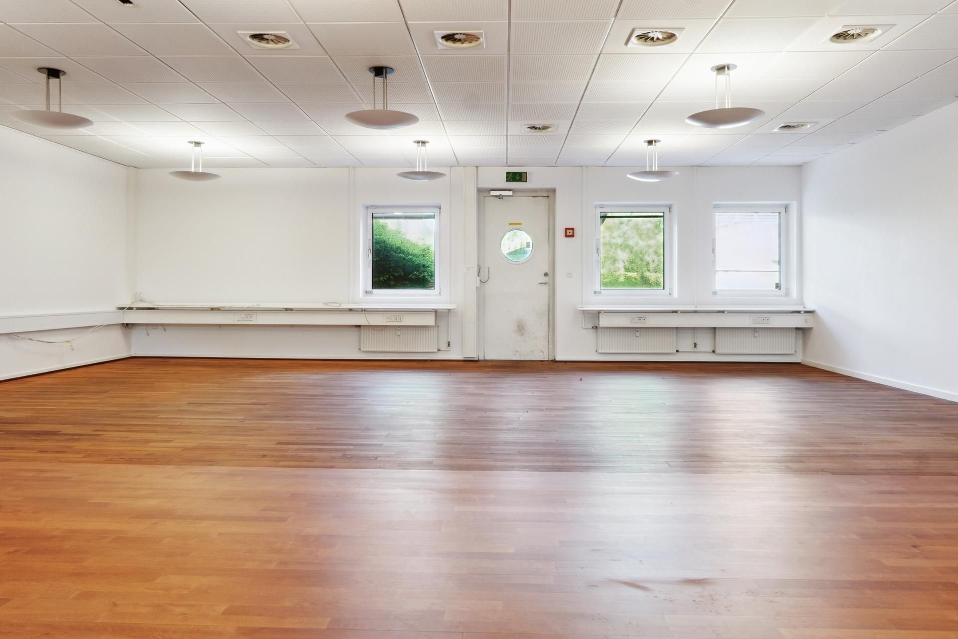 Kontorlokale med hvide vægge, trægulv og fire vinduer samt dør med rundt vindue og lyskroner i loftet