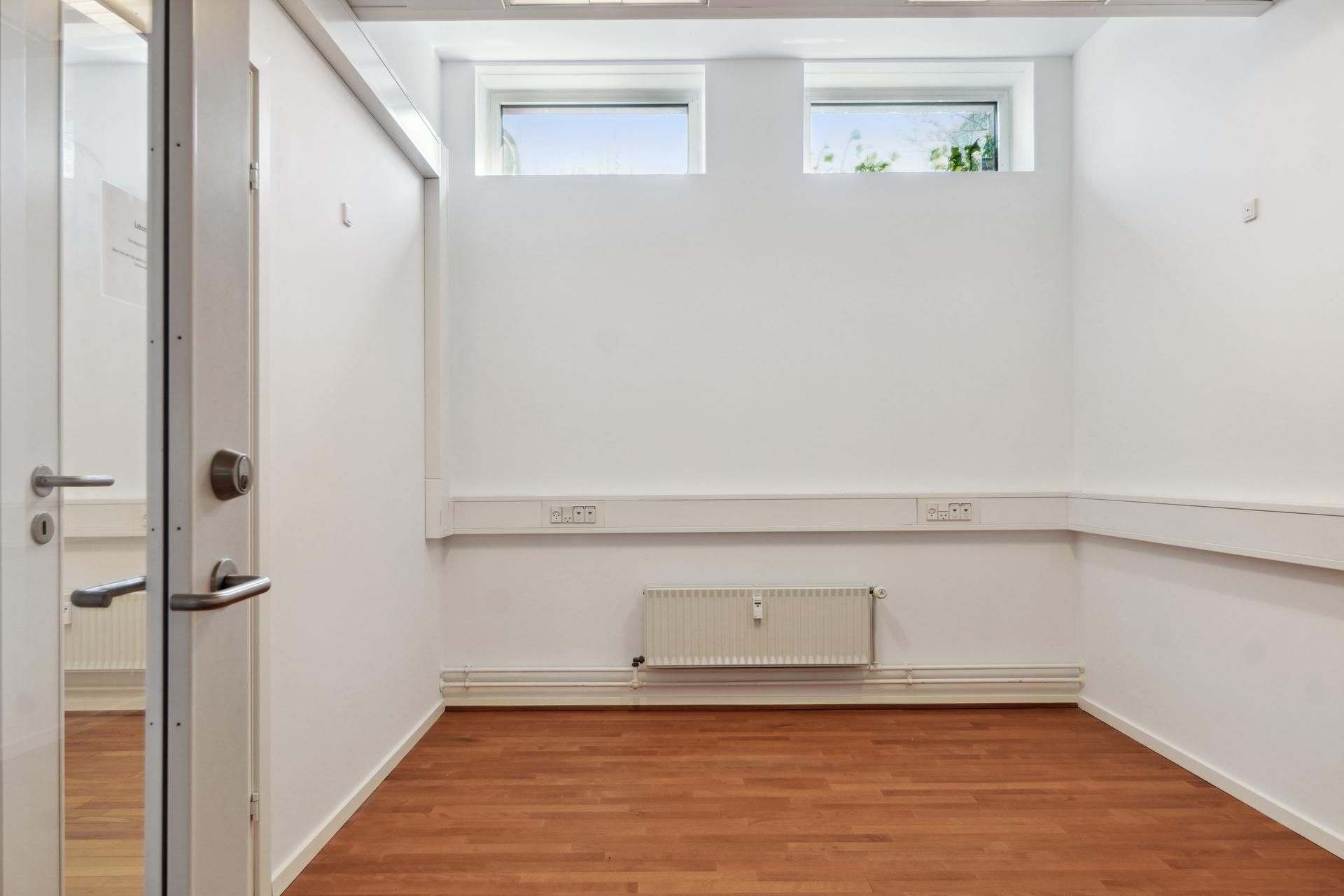 Et tomt kontorlokale med hvide vægge og et åbent glasdør, hvor der er radiatorer og elektriske stikkontakter i væggen
