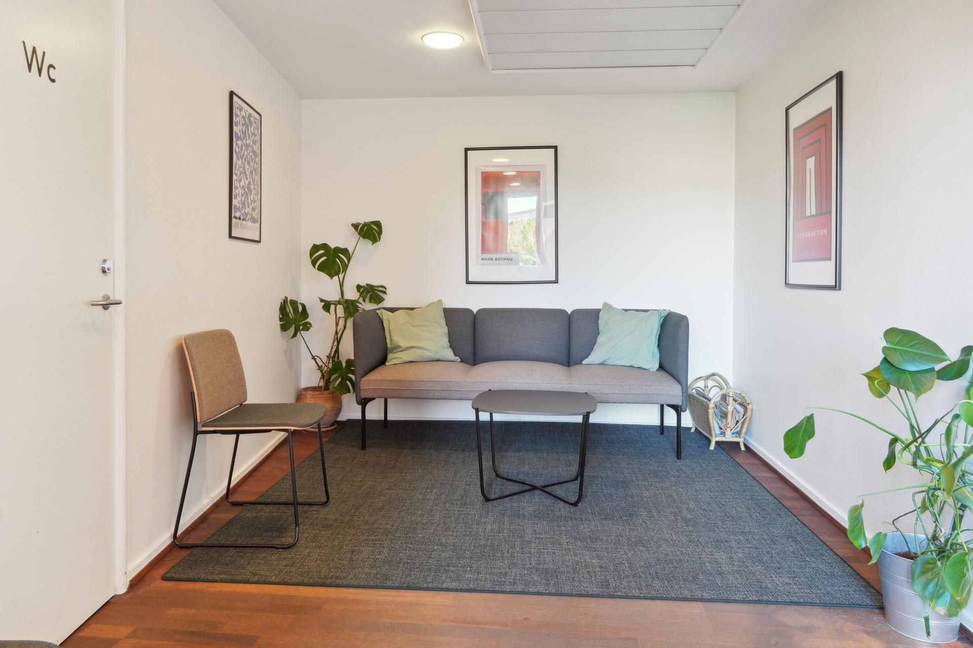 Kontor med et sovesofa, sidebord, plante og stol. Væggene er hvide og der er et wc-mærke på den ene dør.