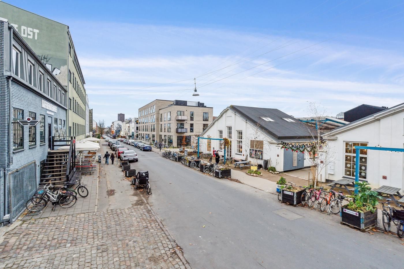 Gade med to sider, hvor der er parkerede biler, cykler og grønne planter i potter, foran huse med forretningsvinduer og