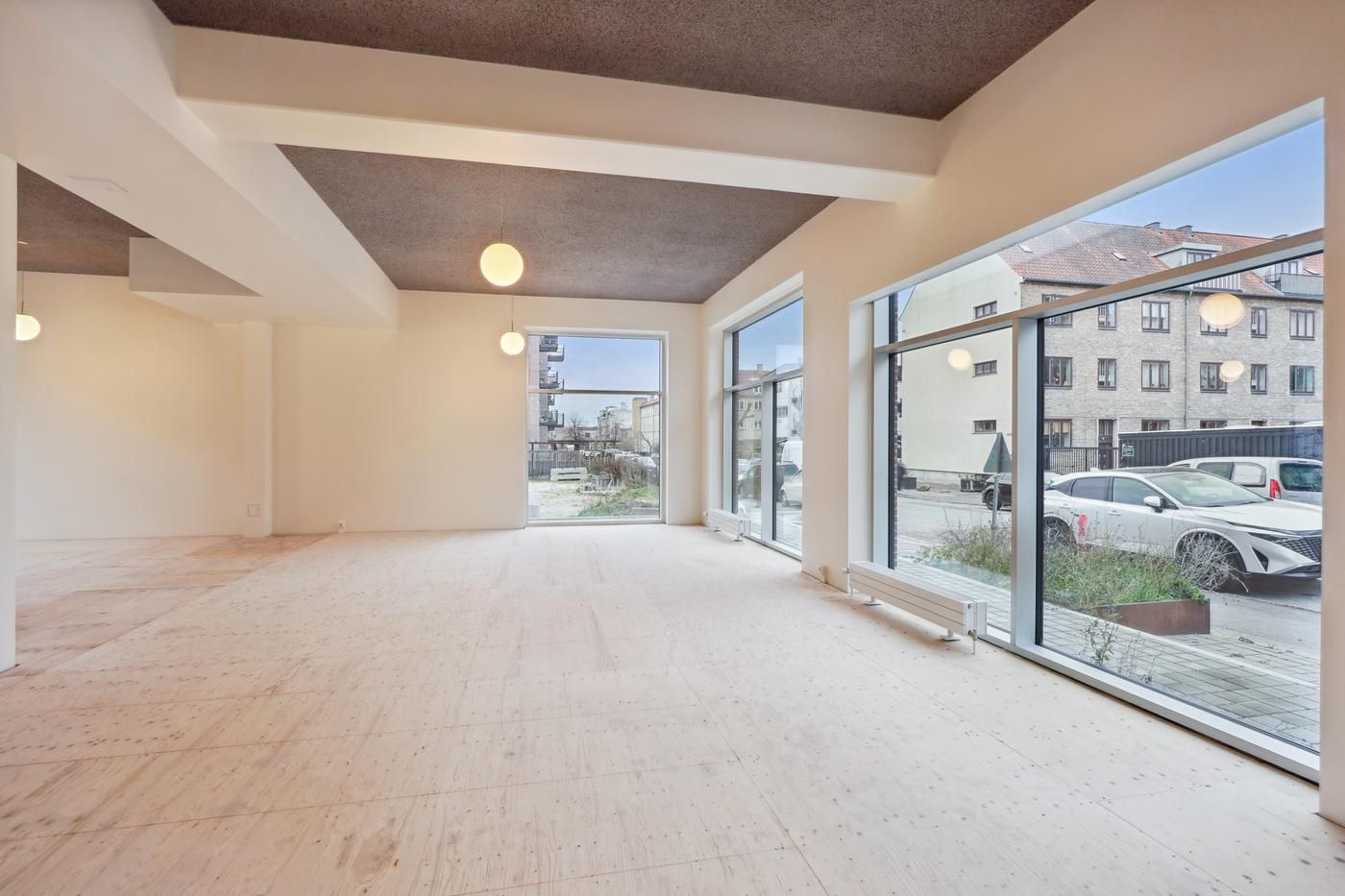 Store lokale med højloft og store vinduer udsat mod en bilparkering med en hvid bil og en bygning i baggrunden