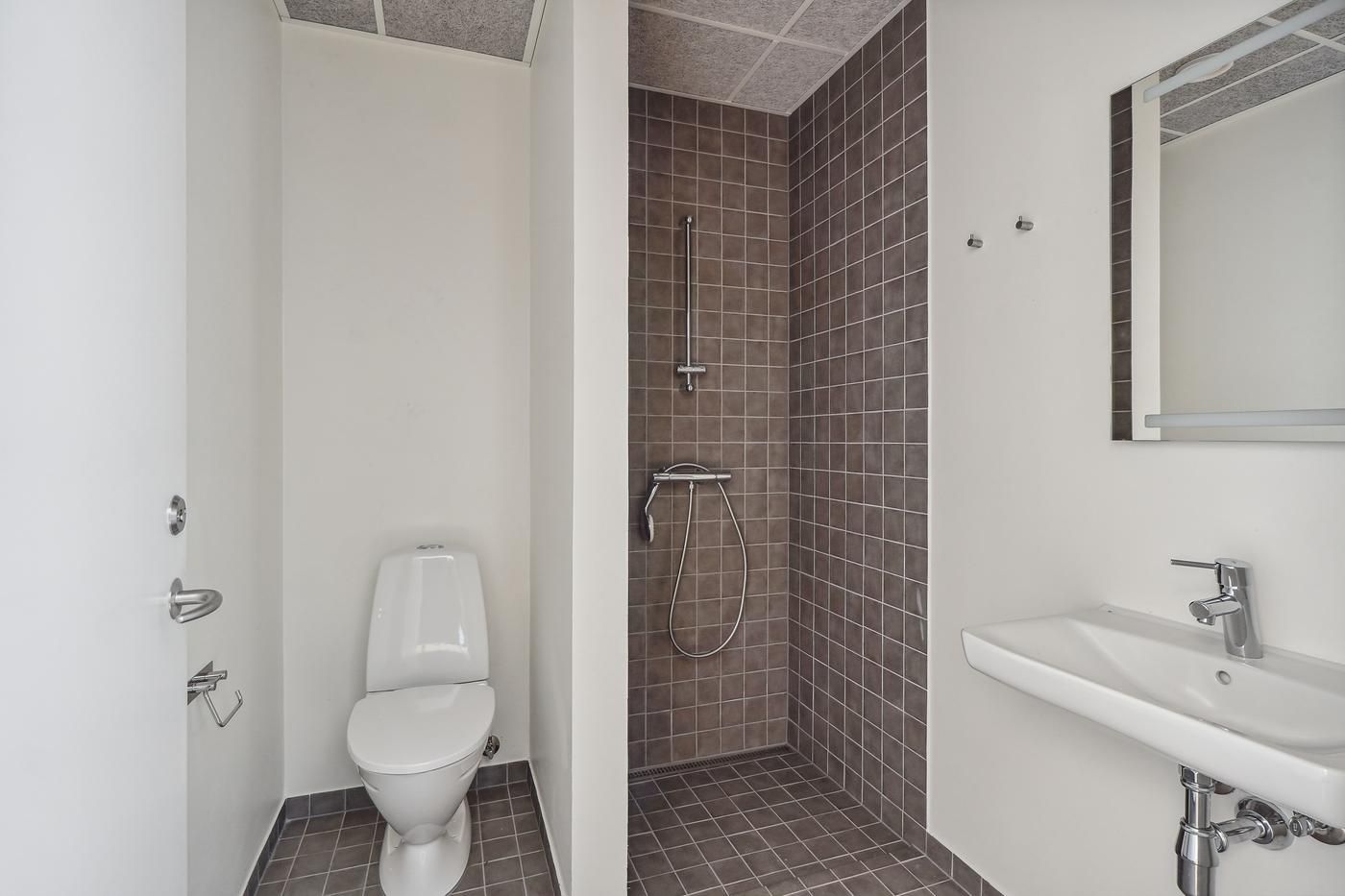 Rent og moderne badeværelse med hvid toilet, lysegrå fliser og rustfrit stål håndtag. Lys og luftig indretning med en stor