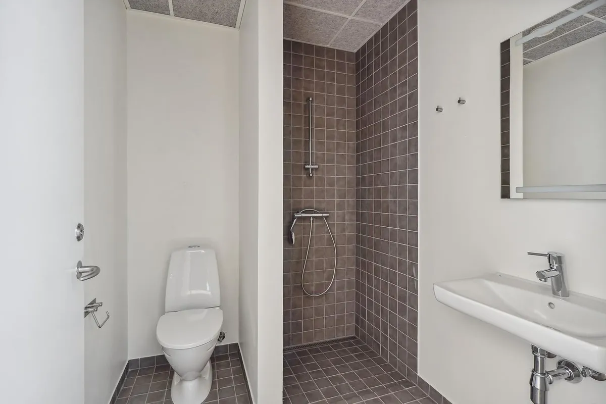 Rent og moderne badeværelse med hvid toilet, lysegrå fliser og rustfrit stål håndtag. Lys og luftig indretning med en stor