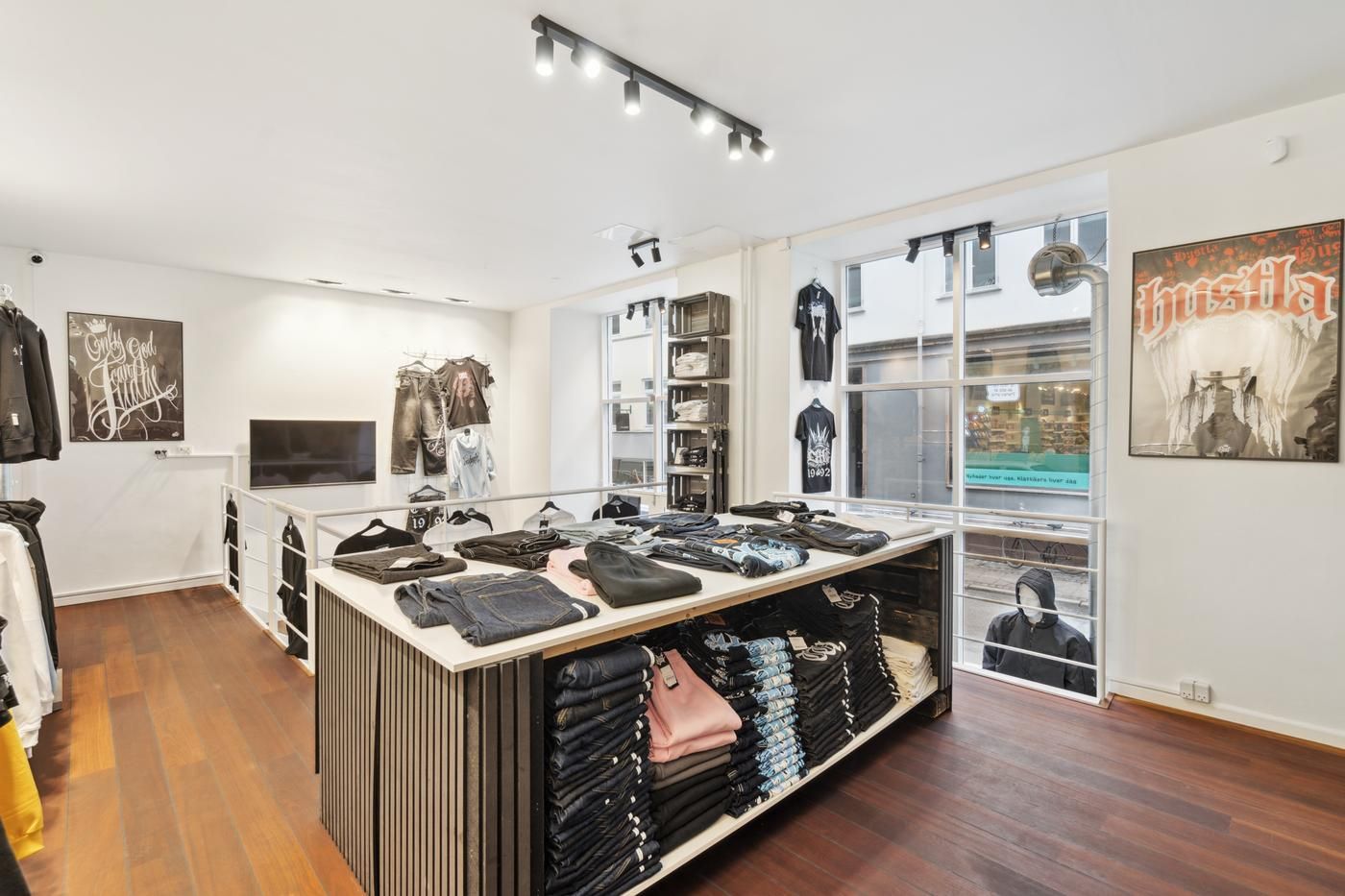 Indbydende butik med tøj på lager, mærket Bushila. Tøjet er udsat på snedkerborde og vægskabe. Plakater hænger på væggen.